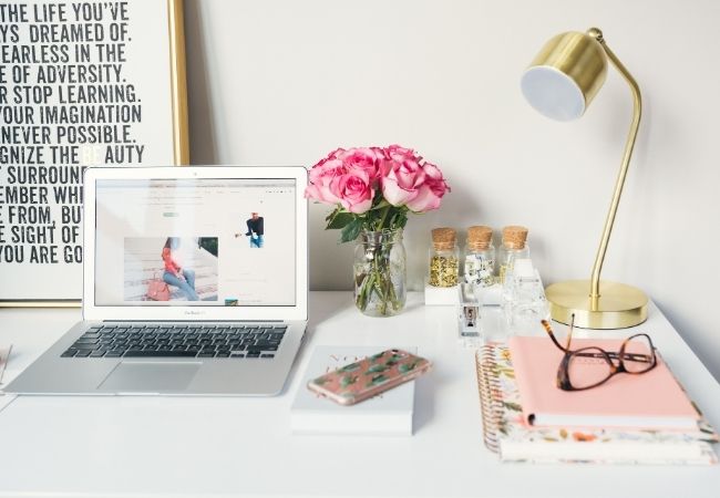 Mesa de home office com notebook, luminária, vaso com flores, caderno, óculos e outros objetos.